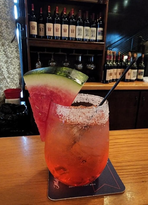 A refreshing cocktail with a salted rim, garnished with a slice of watermelon, sits on a wooden bar top.